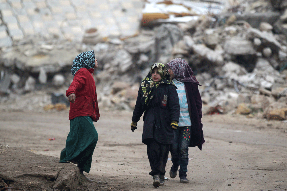 فتيات يسرن على الأقدام بجوار مبان متضررة في ناحية الراعي بمحافظة إدلب